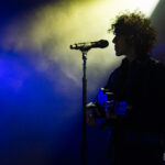 Silhouette of the musician LP playing guitar beside a microphone stand, lit by dramatic blue and yellow stage lights.