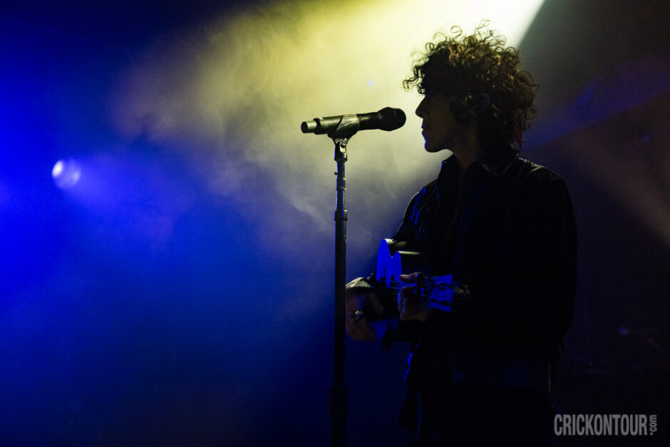 Silhouette of the musician LP playing guitar beside a microphone stand, lit by dramatic blue and yellow stage lights.