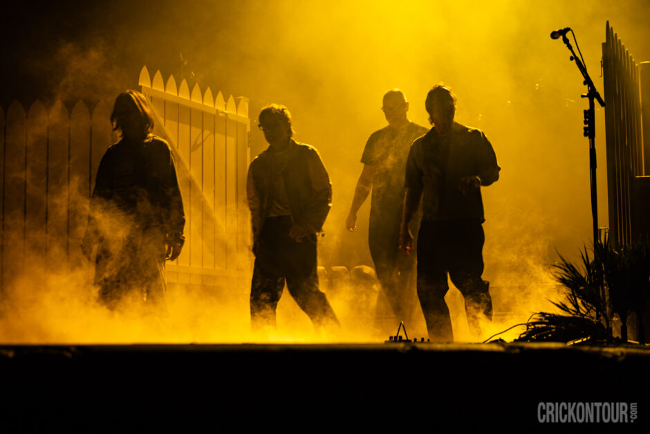Four band members of the band Weezer silhouetted against glowing yellow stage haze, walking past a white picket fence with a microphone stand nearby.