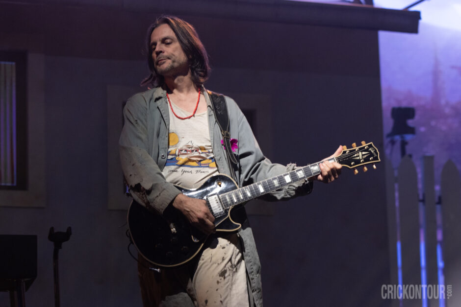 A long-haired Brian Bell of the band Weezer plays his black Gibson Les Paul on stage, wearing a graphic tank top and worn jacket under purple lighting.