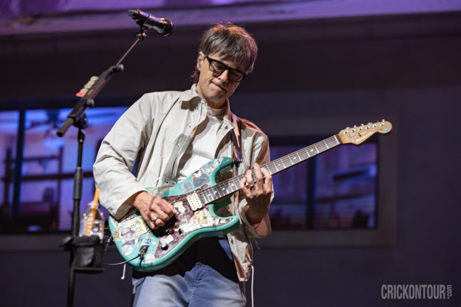 Lead singer and guitarist Rivers Cuomo of Weezer wearing a light jacket plays a sticker-covered teal electric guitar under stage lighting, gazing down at the fretboard.