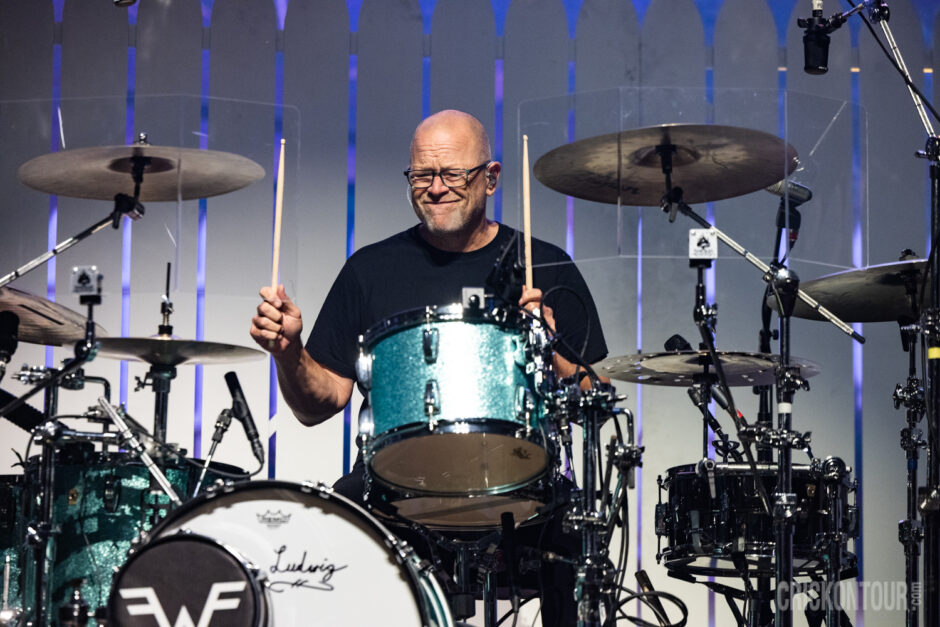 Drummer Patrick Wilson of Weezer plays a teal sparkle drum kit onstage, sticks raised, smiling, in front of a blue-lit backdrop.