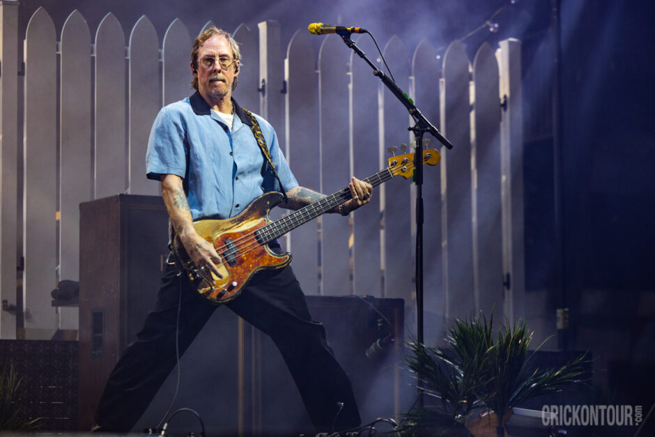 Bassist Scott Shriner of Weezer plays a worn sunburst bass on stage, wearing a light blue shirt and glasses, before a white picket fence backdrop.