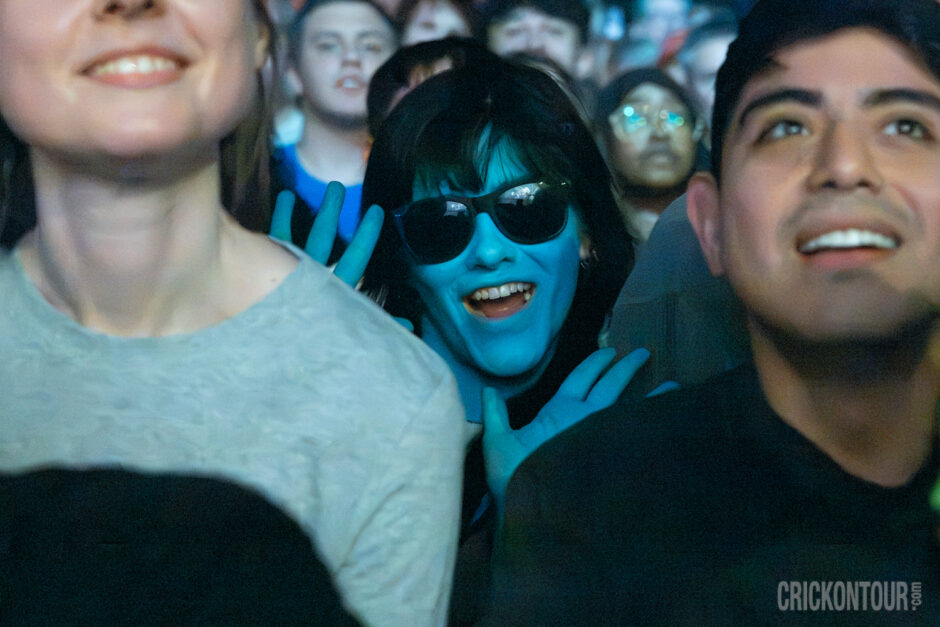 Enthusiastic Weezer fan in a concert crowd bathed in blue paint, wearing sunglasses and grinning while holding hands up in a Weezer sign..