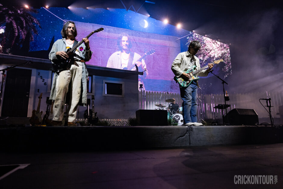 Weezer performs on a stage designed like a suburban house at night, with two guitarists in front, a drummer at center, and a large starry-sky backdrop.