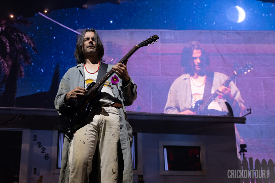 Guitarist Brian Bell of Weezer performing live in front of a suburban house set numbered 2226, wearing a t-shirt with a pink flower and red bead necklace, his image mirrored on a big screen.
