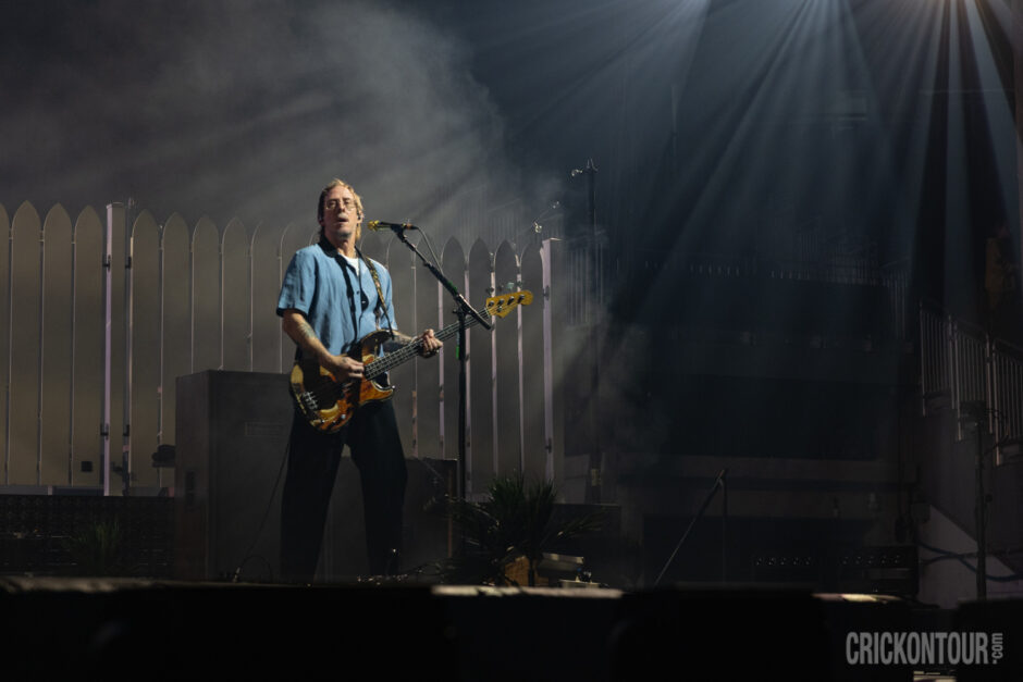 Bassist Scott Shriner of Weezer plays a worn bass guitar and sings into a mic on a smoky, dramatically lit stage with a white picket fence backdrop.
