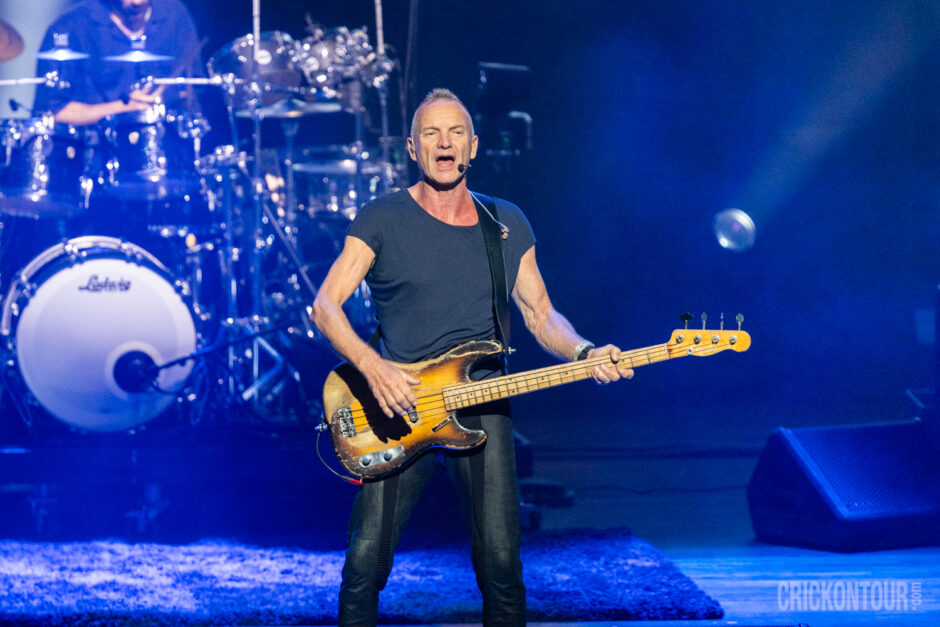 Sting sings while playing a worn sunburst bass guitar on stage, a drum kit with Ludwig bass drum behind him under blue lighting.